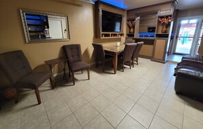 A living room with a dining table, four chairs, a sofa, a TV mounted above the table, and a kitchen area visible through an open door.