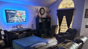 Een moderne slaapkamer met een bed, een massagestoel, een aan de muur bevestigde televisie en een boekenkast.