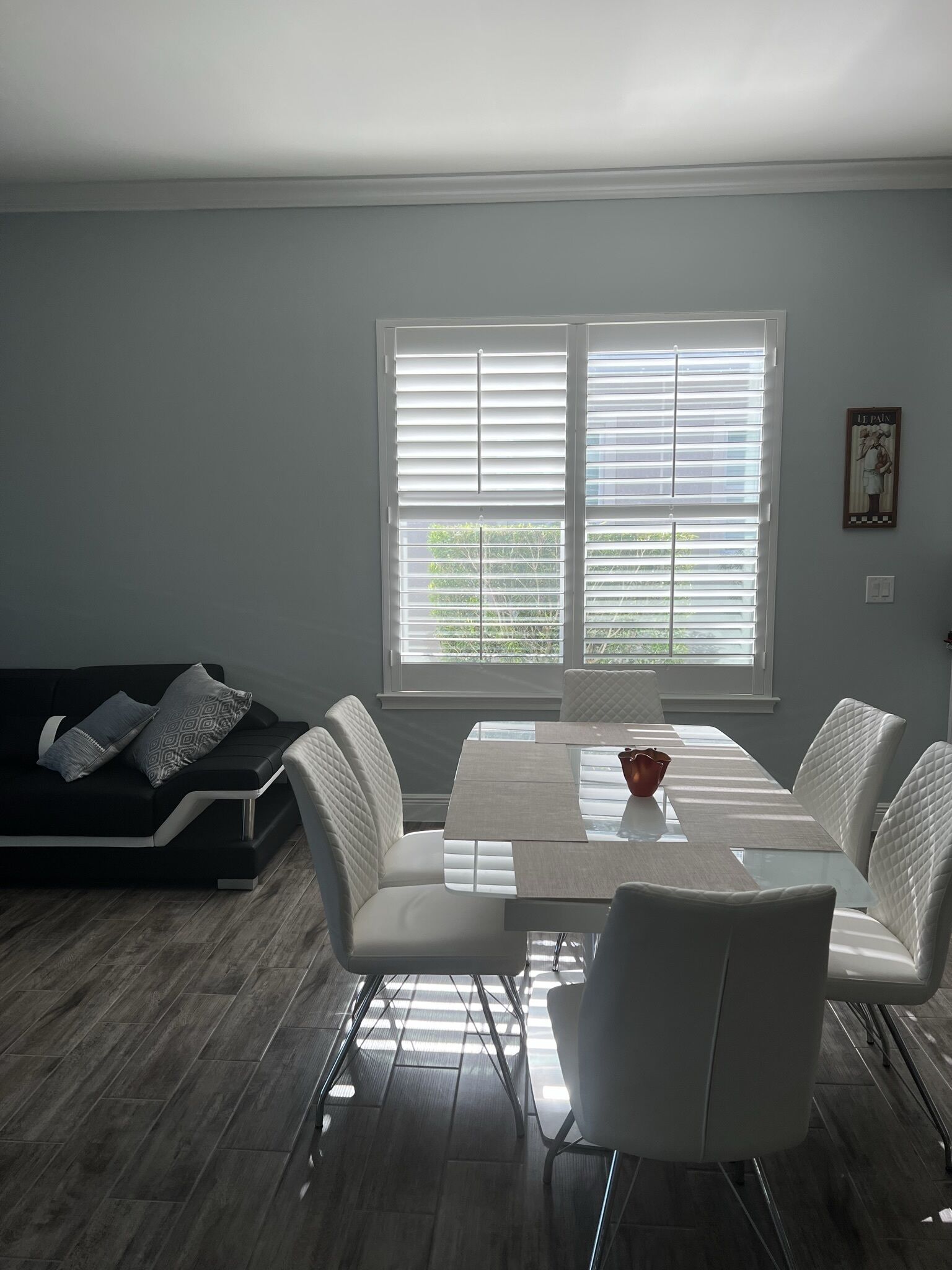 Een moderne woonkamer met een eethoek, met een bank, stoelen, een tafel met glazen blad en een raam met zonwering.