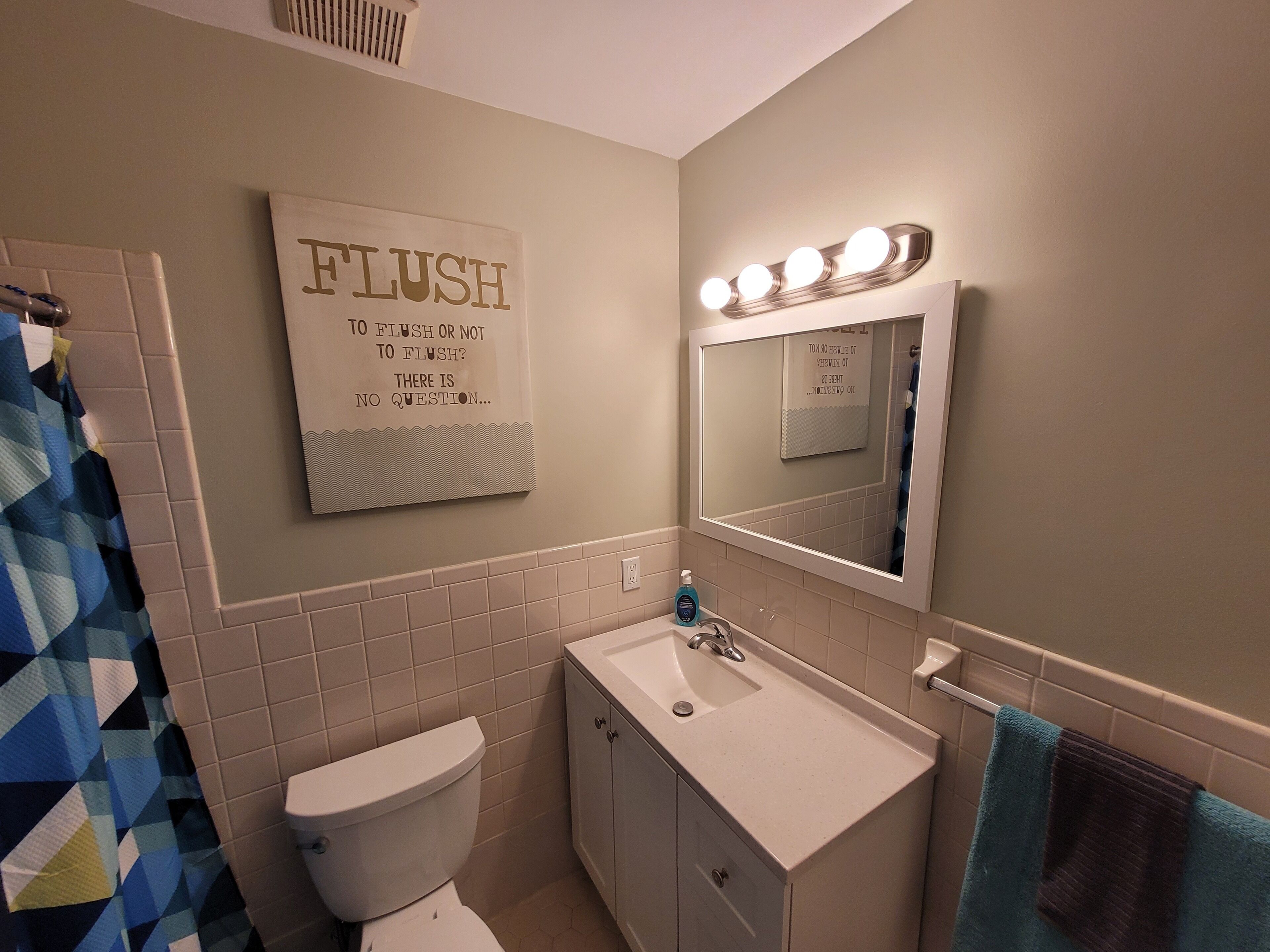 A bathroom with a toilet, sink, vanity, and a mirror with lights. There's a sign on the wall and a towel rack.