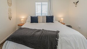 A bedroom with a large bed, two bedside tables with lamps, a window with blinds, and wall decorations including a dreamcatcher and a skull.