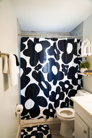 A bathroom with a shower curtain featuring a black and white abstract pattern, a toilet, a sink, and a small shelf with toiletries. - Groovy Getaway - 70s themed house! (Phoenix)