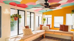 Ein modernes Wohnzimmer mit farbigem Himmel, einem Deckenventilator, einer Bank mit Kissen und hängenden Pflanzen.