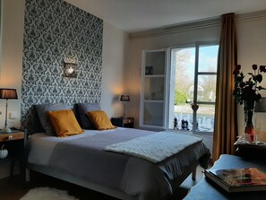 Ein Schlafzimmer mit einem Bett, einem Nachttisch, einer Lampe, einem Fenster mit Vorhängen und einer Vase mit Blumen.