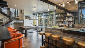 A modern bar area with a pool table, bar stools, and a variety of bottles.