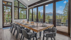 A dining area with a long table set for six, surrounded by chairs, large windows offering a mountain view, and a deck with outdoor seating. - Hot Tub + Ski Shuttle | Vickery's Vantage (Breckenridge)