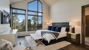 Une chambre avec un grand lit, deux lampes de chevet, un bureau et une chaise. De grandes fenêtres offrent une vue sur les montagnes.