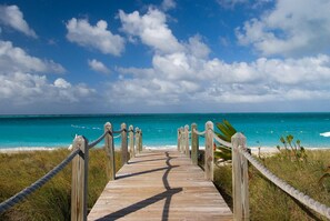 En träpromenad som leder till en strand med turkosblått vatten och en klar himmel.