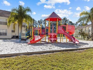 A playground with slides and a climbing structure, surrounded by sand and grass, with buildings in the background.