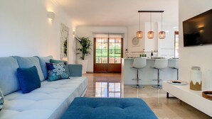 Een moderne woonkamer met een witte bank, een blauwe poef en een keukenhoek met barkrukken en een televisie.