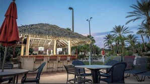 An outdoor dining area with thatched-roof seating and tables, surrounded by palm trees. - 3 bedroom villa (Sleeps 8) in a tropical Tuscan style resort near Disney (Davenport)