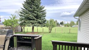 A deck with a grill and outdoor furniture, overlooking a grassy yard with trees and a fence. - Close to Glacier Park, Flathead Lake, Whitefish Mountain Resort (Kalispell)