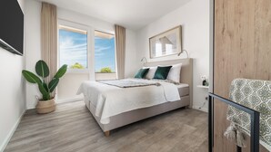 A bedroom with a bed, a large window, a plant, and a chair. - "BeachLoungeTwo" vacation apartment on the beach with sun terrace, sauna & sea view (Olpenitz)