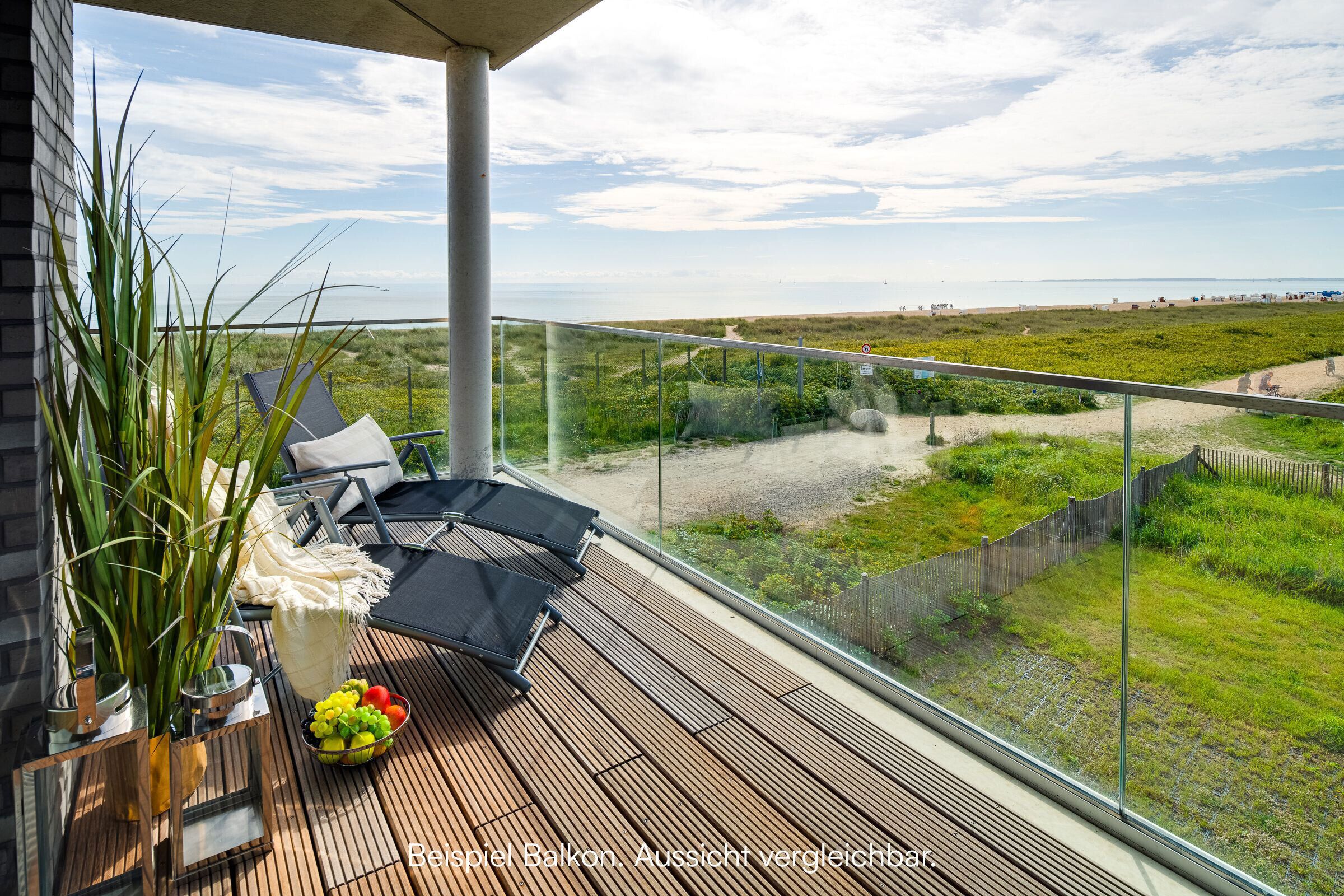 A balcony with a view of the beach, a glass railing, and a table with fruits and a vase of plants.