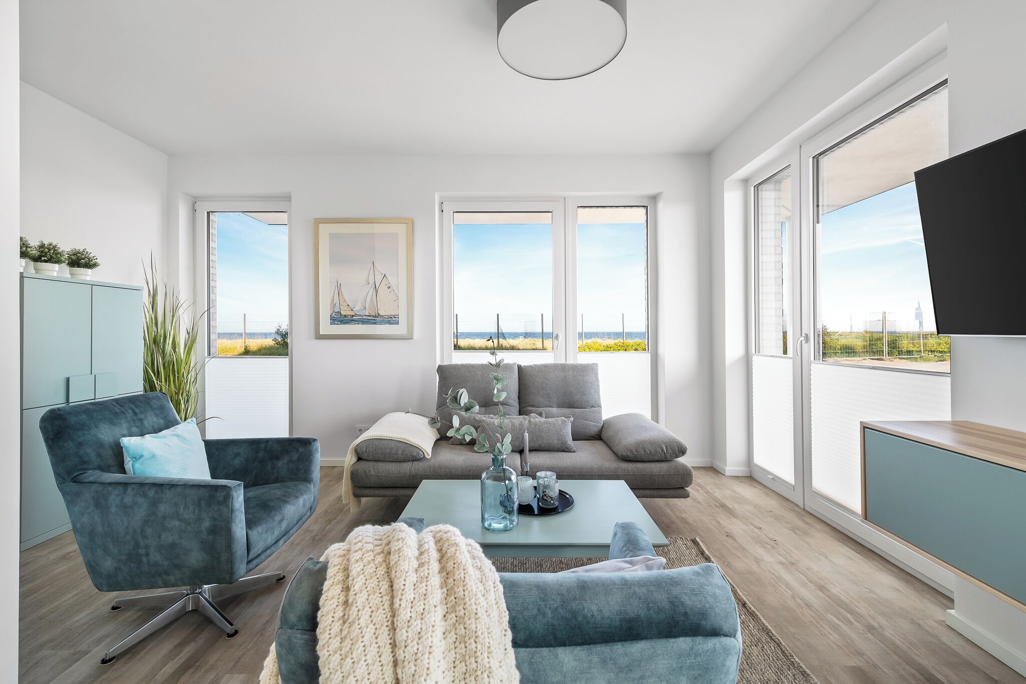 A modern living room with a grey sofa, a blue armchair, and a coffee table. There are large windows with a view of the beach and a sailboat.