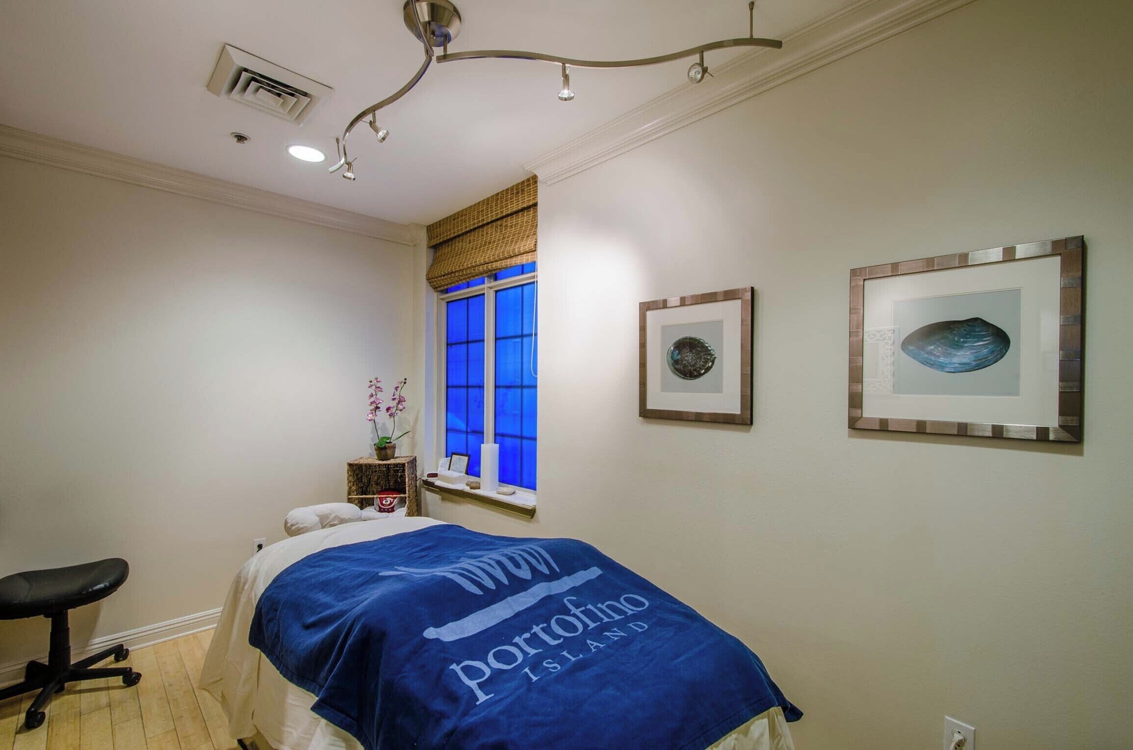 A bedroom with a bed, a chair, a window with blue-tinted glass, and two framed pictures on the wall.