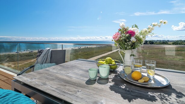 A balcony with a table set with fruits, glasses, and a flower vase, overlooking a beach and ocean. - "BeachViewTwo": Holiday apartment right on the beach with sauna, balcony & Baltic Sea view (Olpenitz)