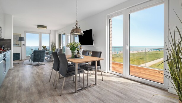 A spacious living area with a wooden dining table, modern chairs, and a large window offering a view of the beach. - "BeachViewFour": Holiday apartment right on the beach with sauna, balcony & Baltic Sea view (Olpenitz)