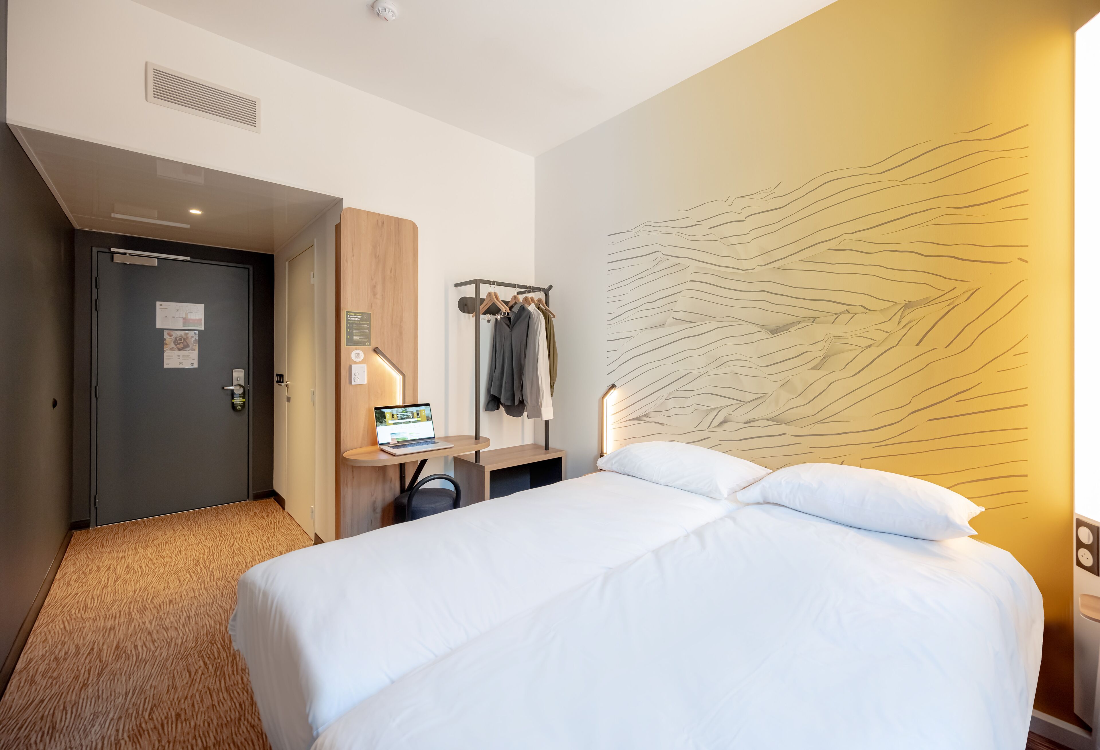 A hotel room with a bed, a desk with a laptop, a coat rack, and a wall with a textured design.