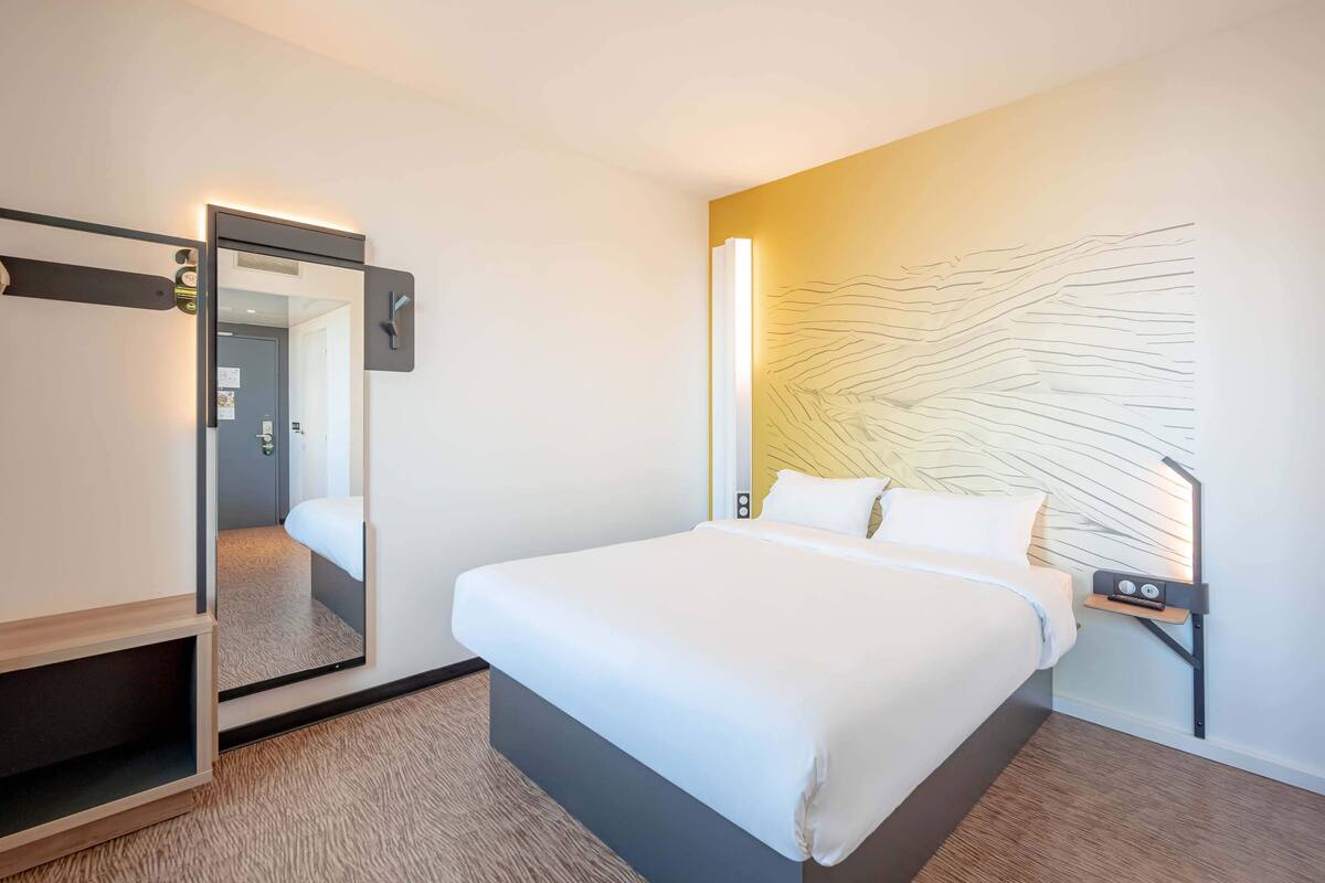 Une chambre d’hôtel moderne dotée d’un grand lit, d’une table de chevet avec une lampe, d’un miroir et d’une œuvre d’art murale originale.