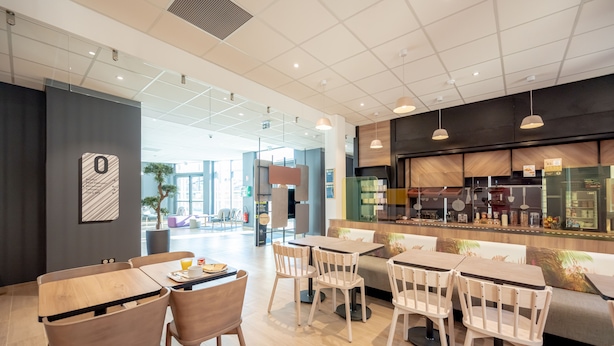 A modern cafe with wooden tables and chairs, a counter with food items, and a large window offering a view of the outside.