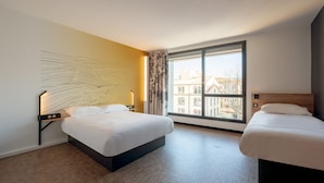 A hotel room with two beds, a large window with a view of buildings, and a modern wall art piece.