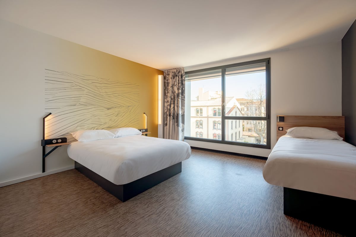 Une chambre d’hôtel avec deux lits, une grande fenêtre donnant sur des immeubles et une œuvre d’art moderne accrochée au mur.