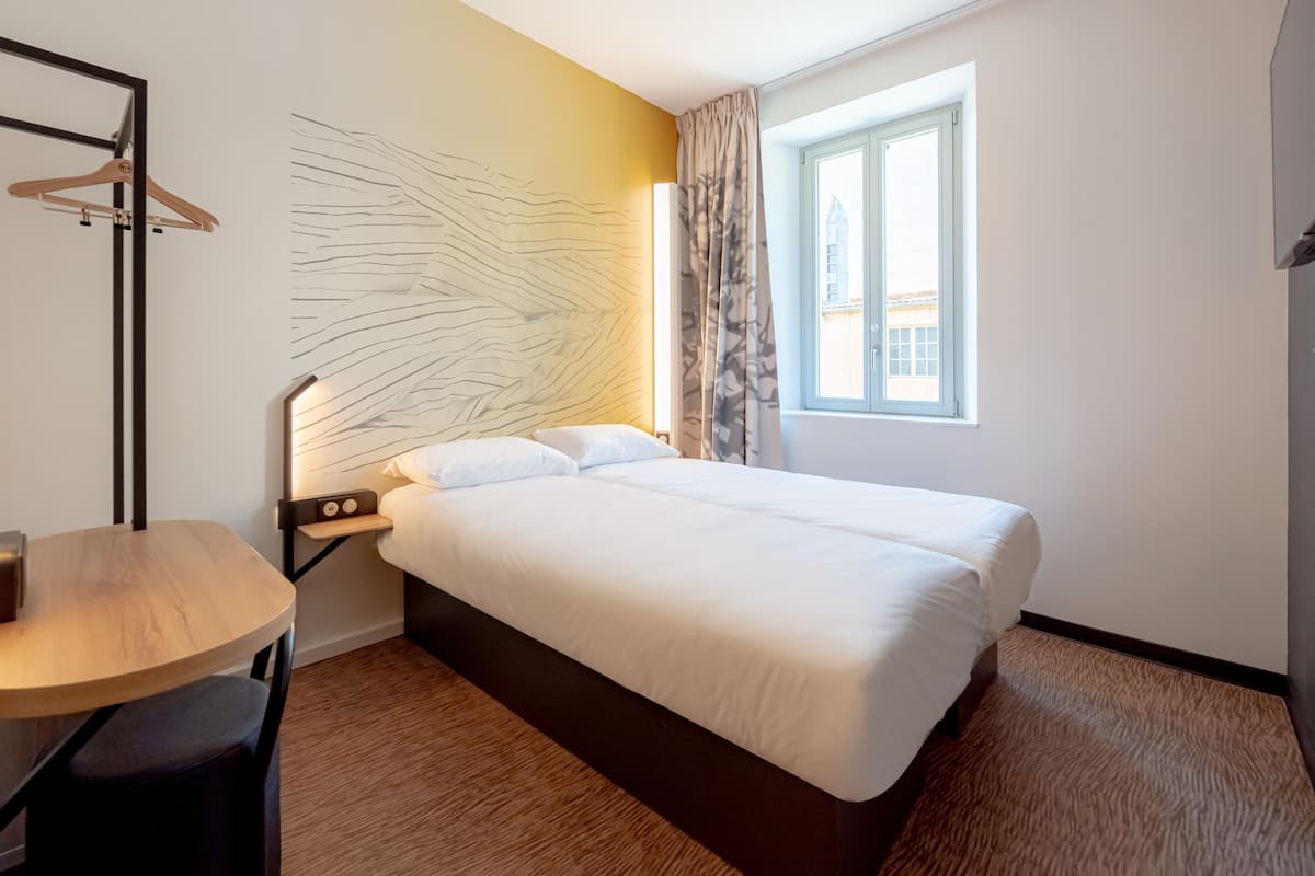 Une chambre d’hôtel comprenant un lit, une table de chevet, une lampe, un porte-manteau mural et une fenêtre donnant sur des immeubles.