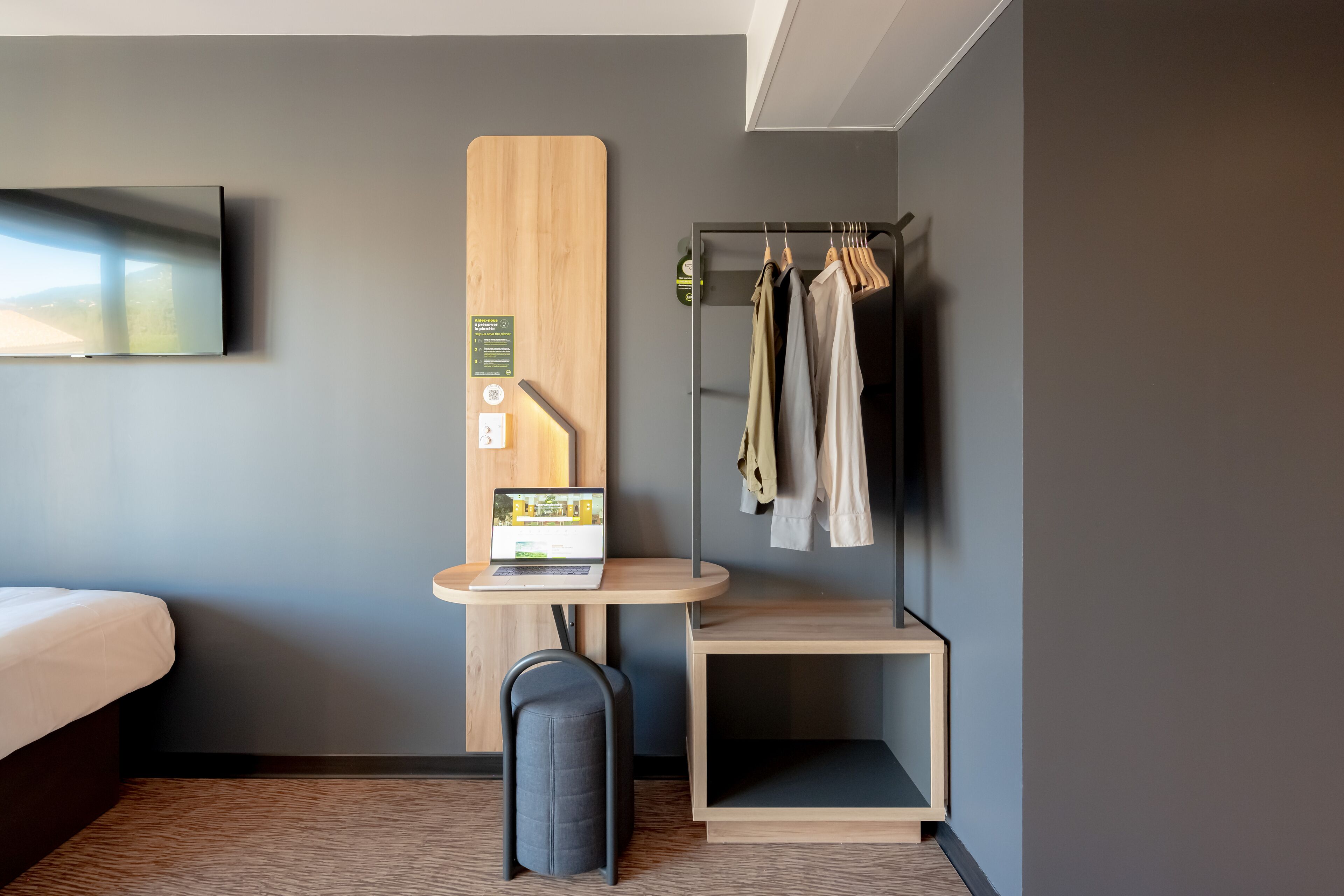 Une chambre d’hôtel moderne équipée d’un lit, d’un bureau avec un ordinateur portable, d’un porte-manteau avec des vêtements et d’un petit coin salon.