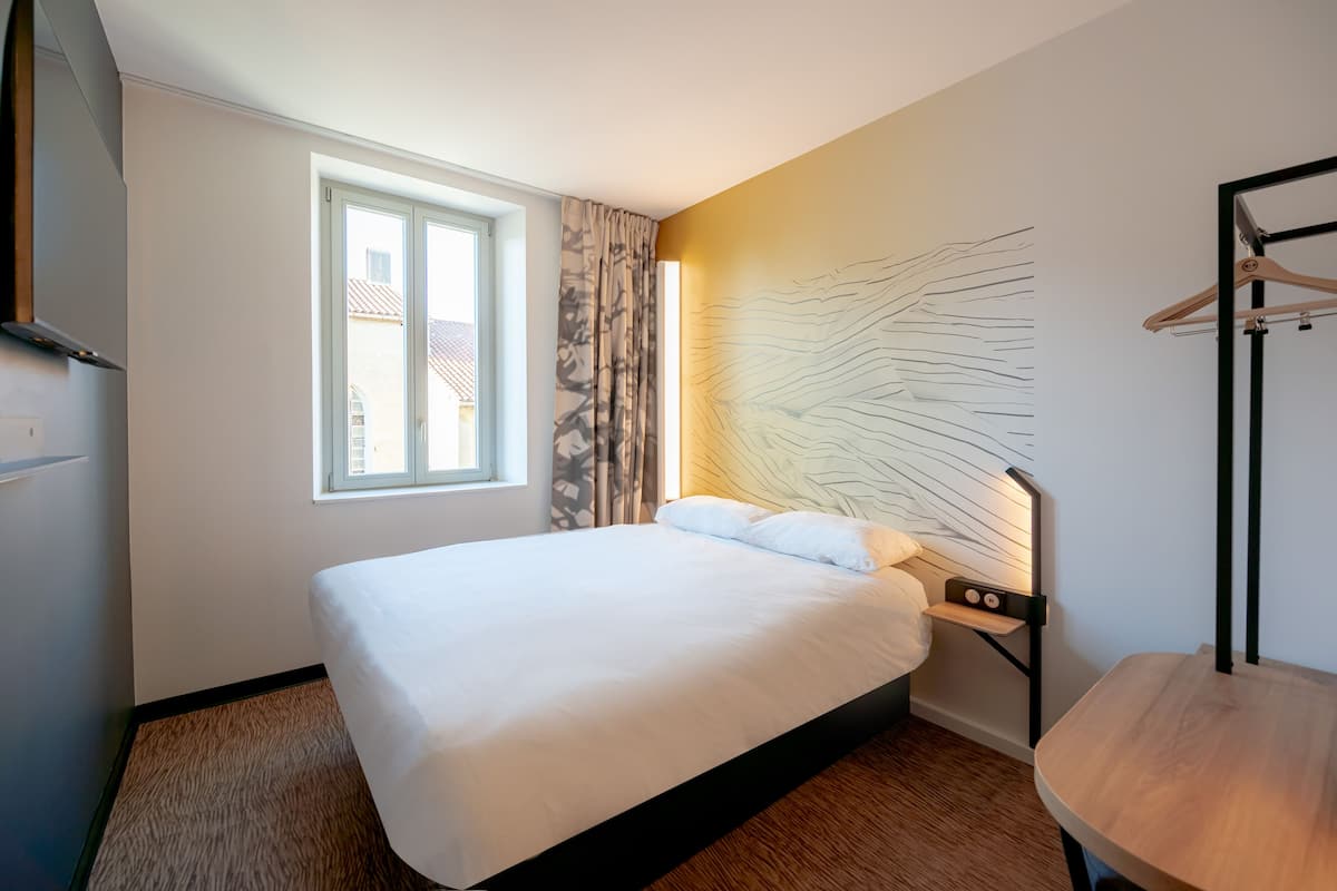 Une chambre d’hôtel avec un grand lit, une table de chevet, une lampe fixée au mur, une fenêtre avec des rideaux et un porte-manteau.