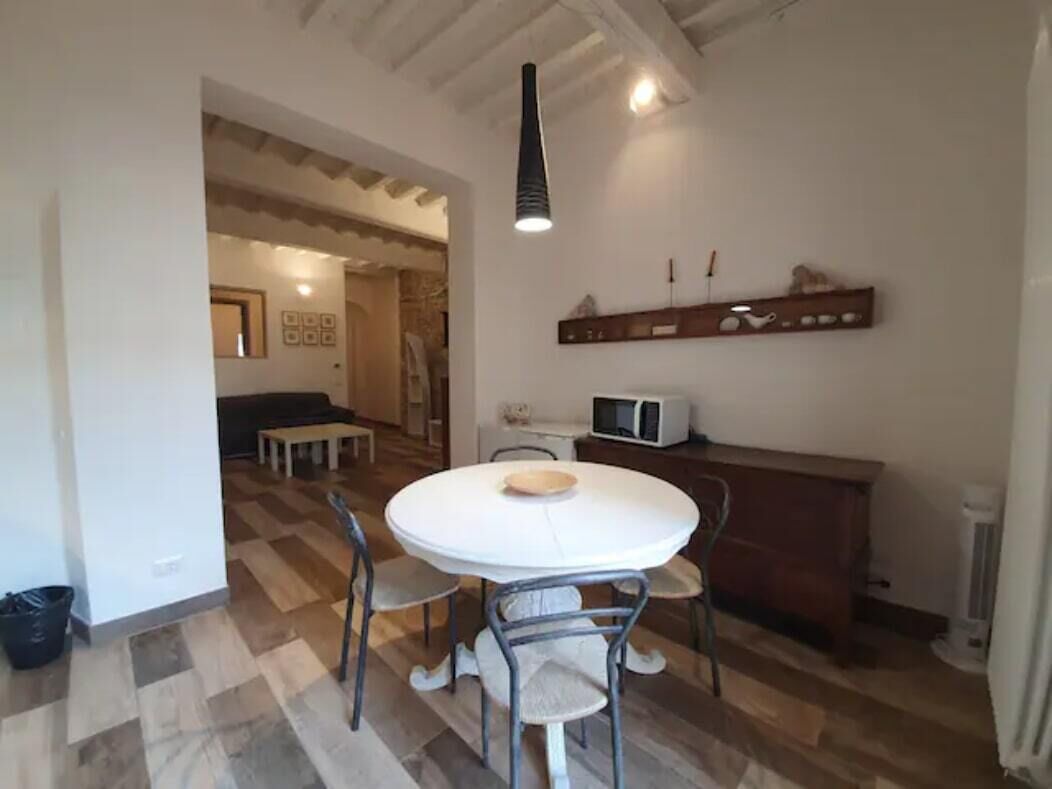 Uma área de refeições com uma mesa redonda, quatro cadeiras, um candeeiro pendente e um micro-ondas em uma bancada.