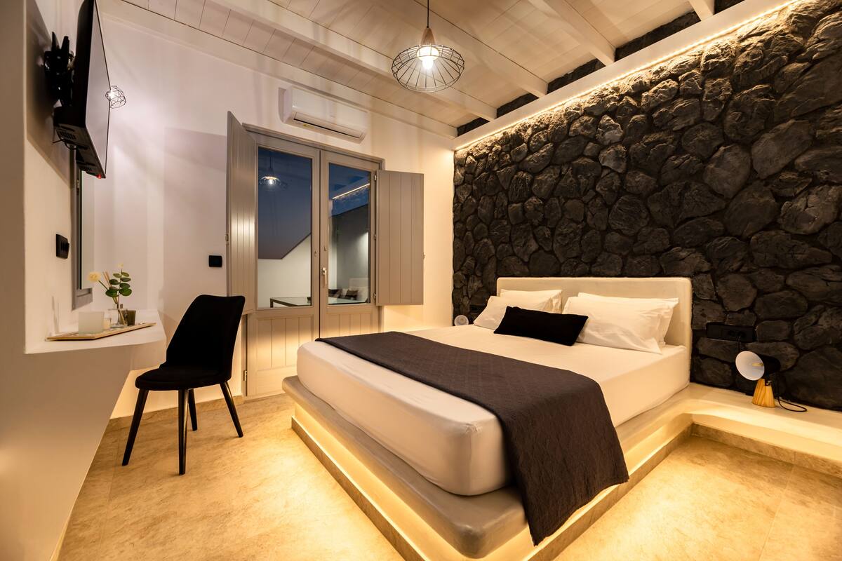 Une chambre moderne avec un mur d’accent en pierre, un grand lit, un fauteuil et un bureau avec un ordinateur portable.