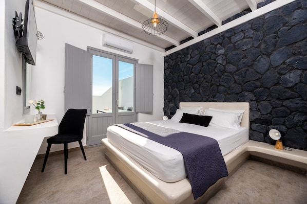 A bedroom with a stone wall, a bed with white bedding, a black chair, and a desk with a plant. - Filoxenia Luxury Villas (Santorini)