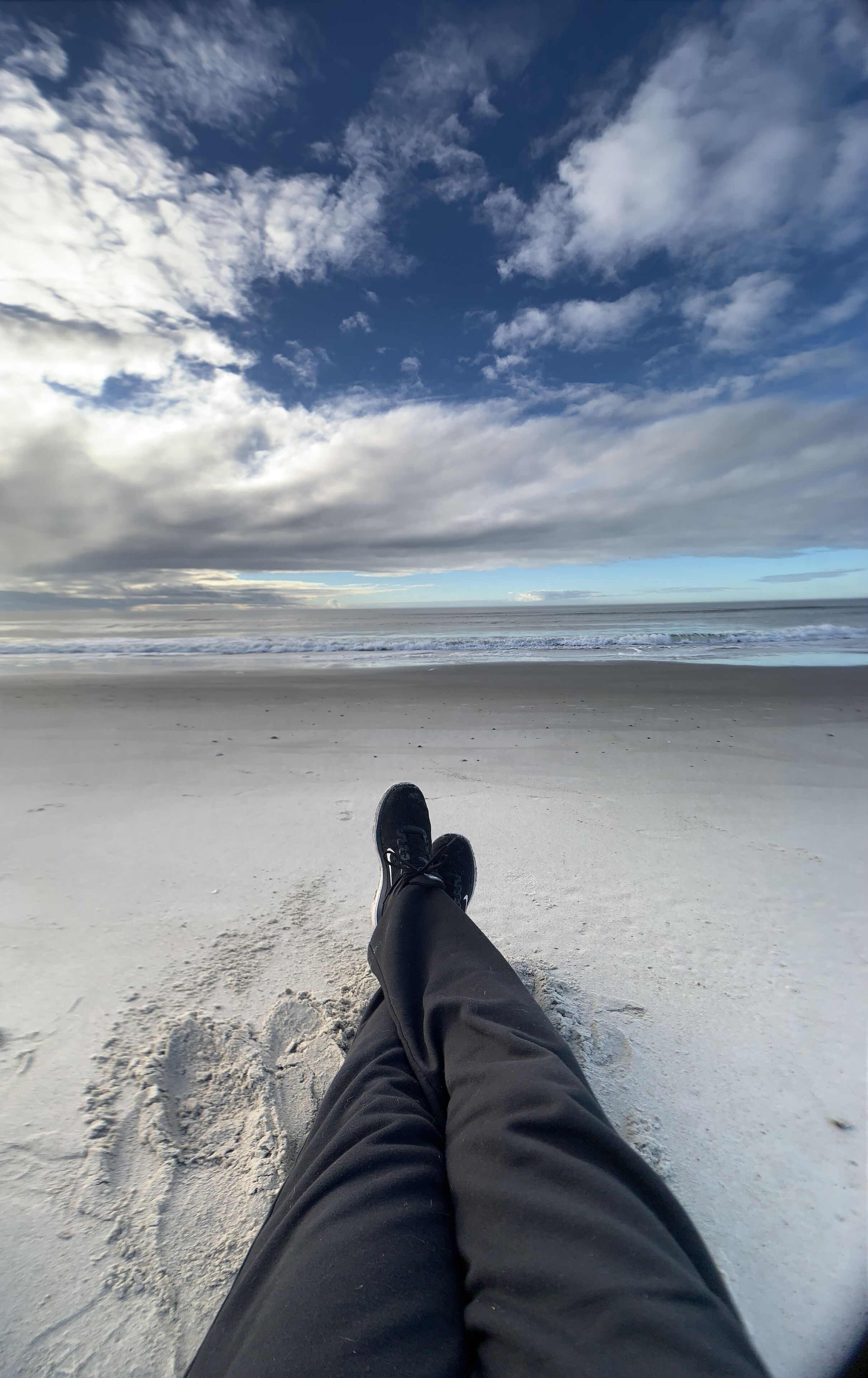 Une vue sur une plage, avec les jambes et les pieds d’une personne, un rivage sablonneux et un ciel nuageux.