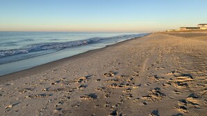 Une plage où l’on voit des empreintes de pas menant à l’eau, un ciel dégagé et des bâtiments en arrière-plan.
