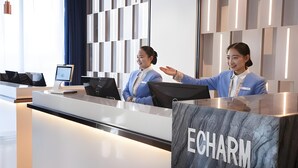 Bancada de recepção de hotel em mármore com a identificação 'ECHARM', duas recepcionistas uniformizadas em azul e design de interiores moderno.
