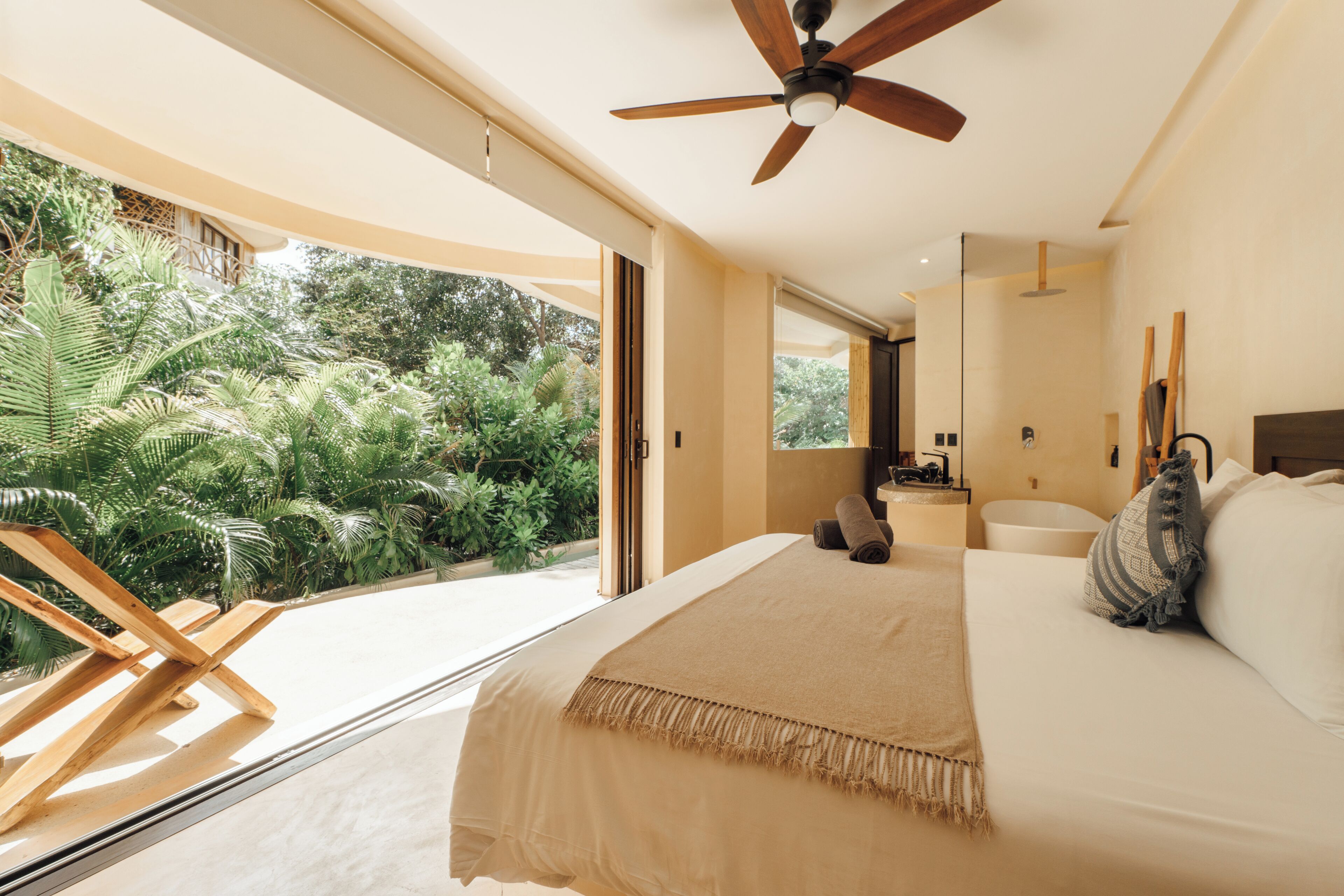Quarto com cama grande, ventoinha no teto, vista de vegetação exuberante e banheiro com banheira.