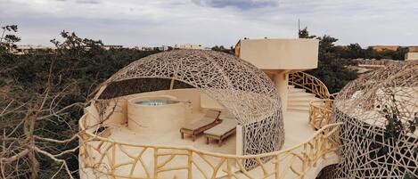 Un edificio moderno de forma abovedada con jacuzzi y zona de estar exterior, rodeado de vegetación natural.