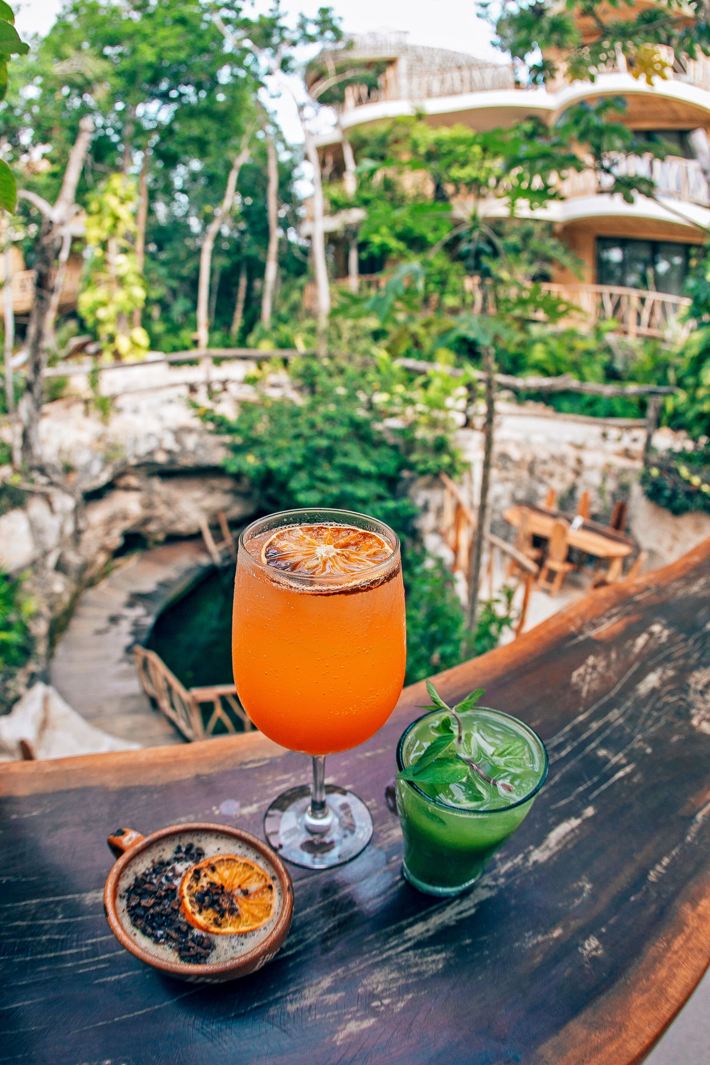 Un vaso de jugo de naranja, una bebida verde y un tazón pequeño con una rodaja de naranja deshidratada sobre una mesa de madera.