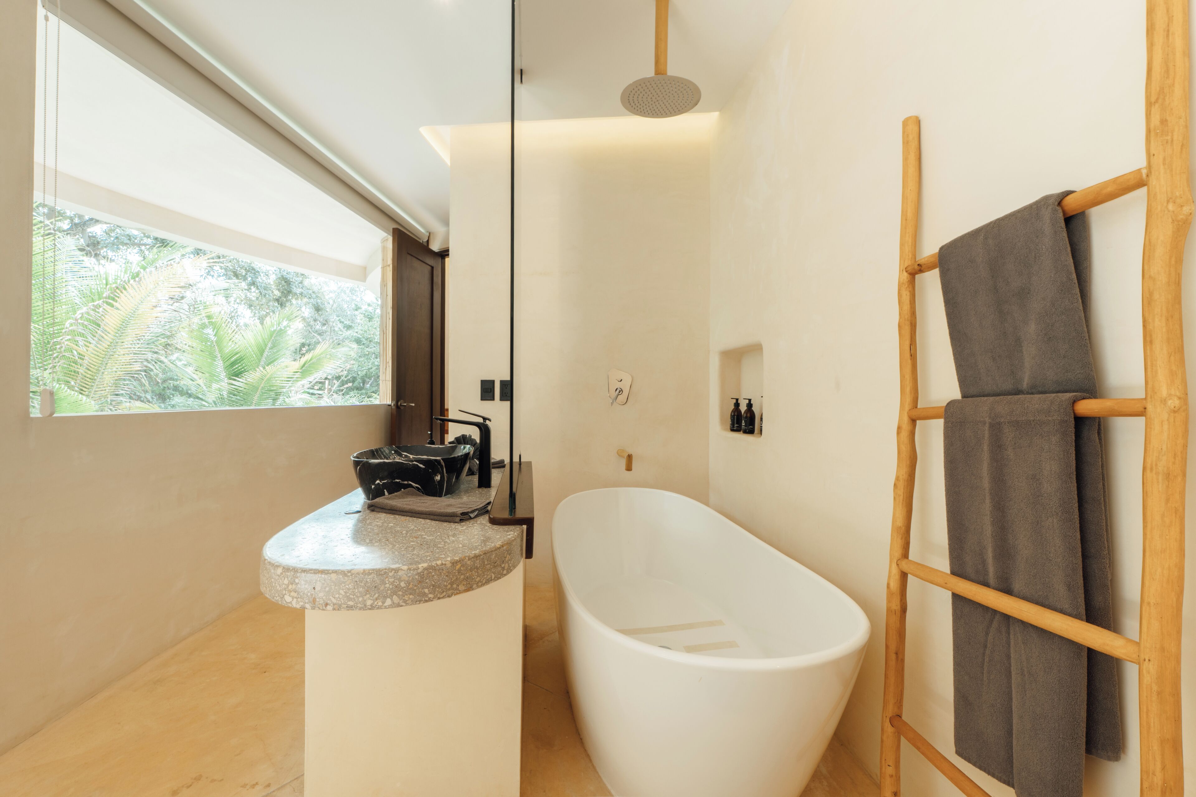 A modern bathroom with a freestanding bathtub, a wooden towel rack, and a window with a view of greenery.