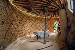 Une pièce circulaire avec un plafond en forme de dôme en bois, un lit et un tronc d’arbre au centre.