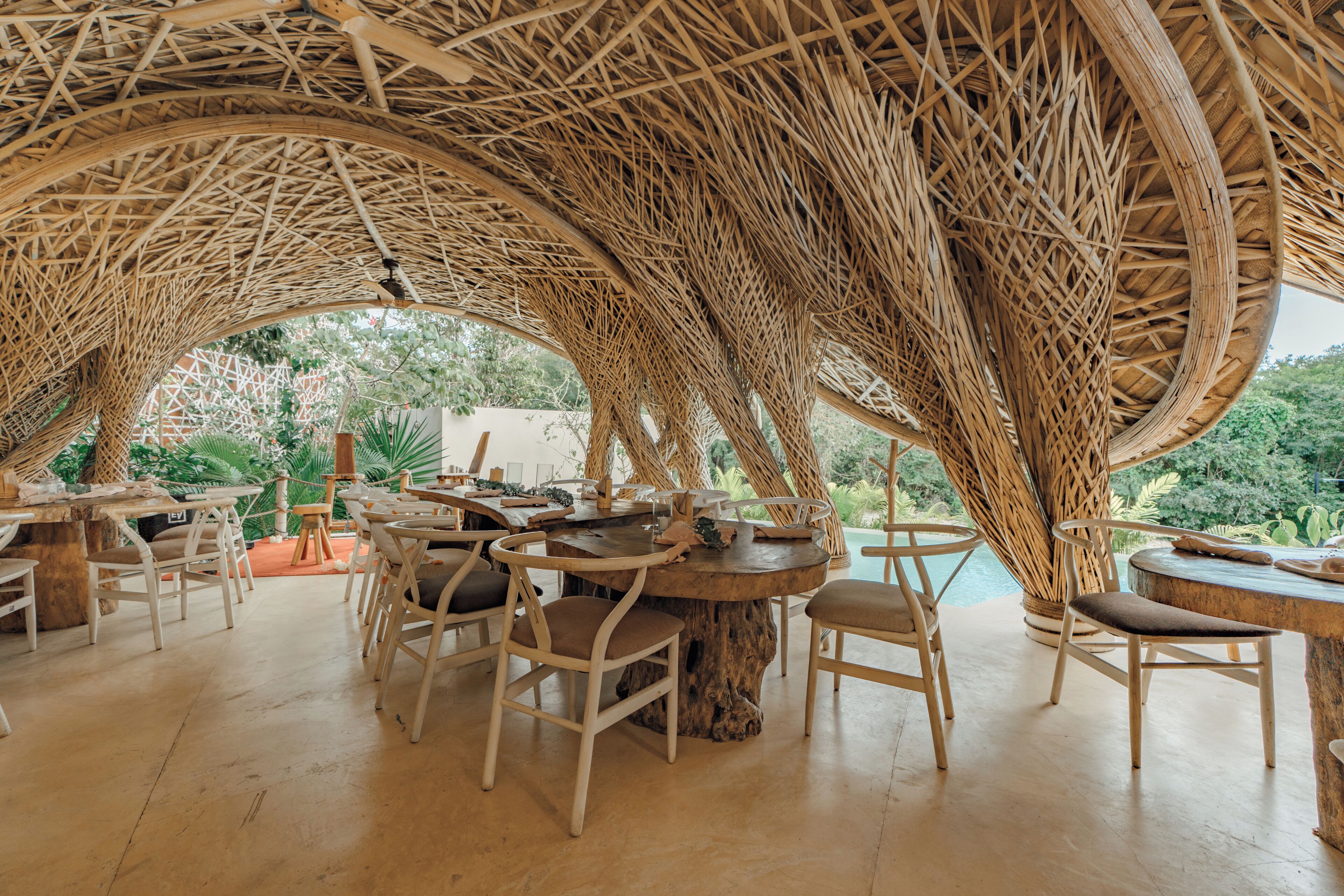 An outdoor dining area with wicker tables and chairs, surrounded by a woven canopy structure.