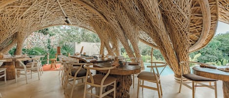 An outdoor dining area with wicker tables and chairs, surrounded by a woven canopy structure.
