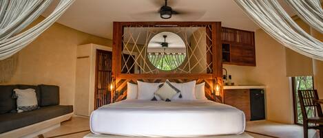 A large bed with a wooden headboard, a ceiling fan, and a view of greenery through a circular window.