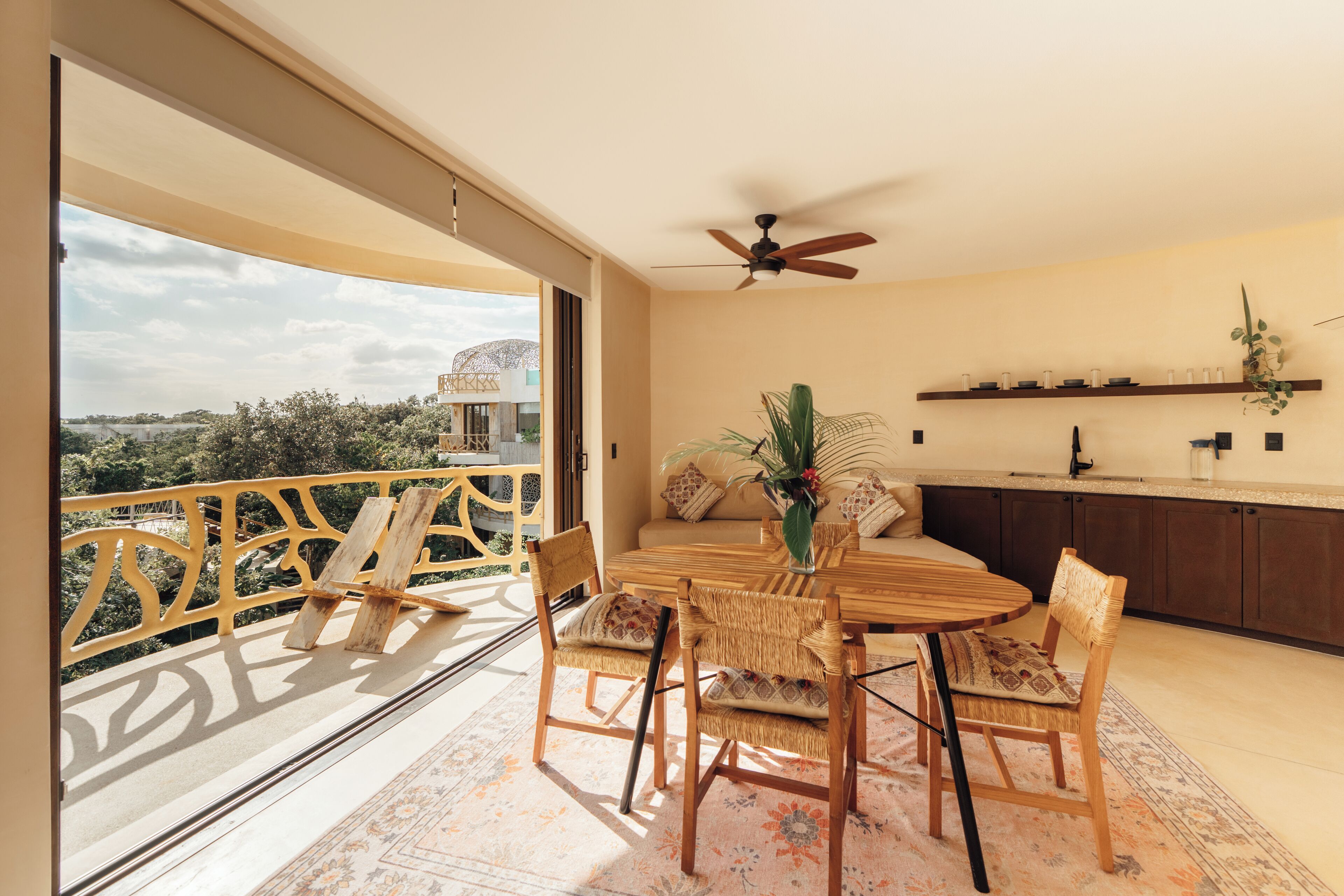 A balcony with a wooden railing, a dining table with chairs, a ceiling fan, and a kitchen area with a shelf above the counter.