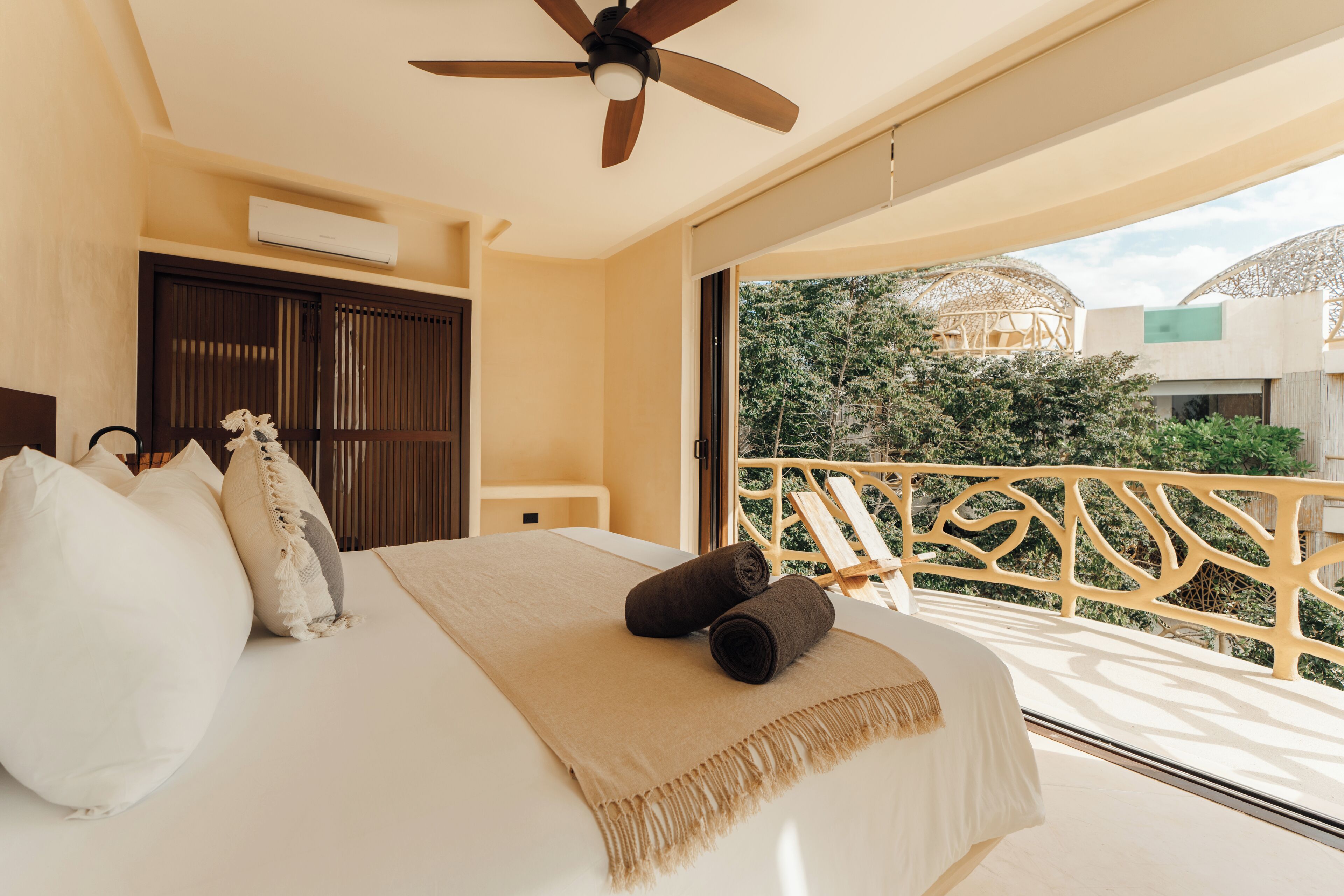 A hotel room with a bed, a ceiling fan, a balcony with a wooden railing, and a view of greenery outside.