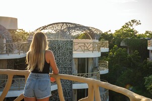 Une personne se tient sur un balcon d’où l’on aperçoit un bâtiment moderne, orné d’un réseau complexe de motifs et dont l’arrière-plan est verdoyant.