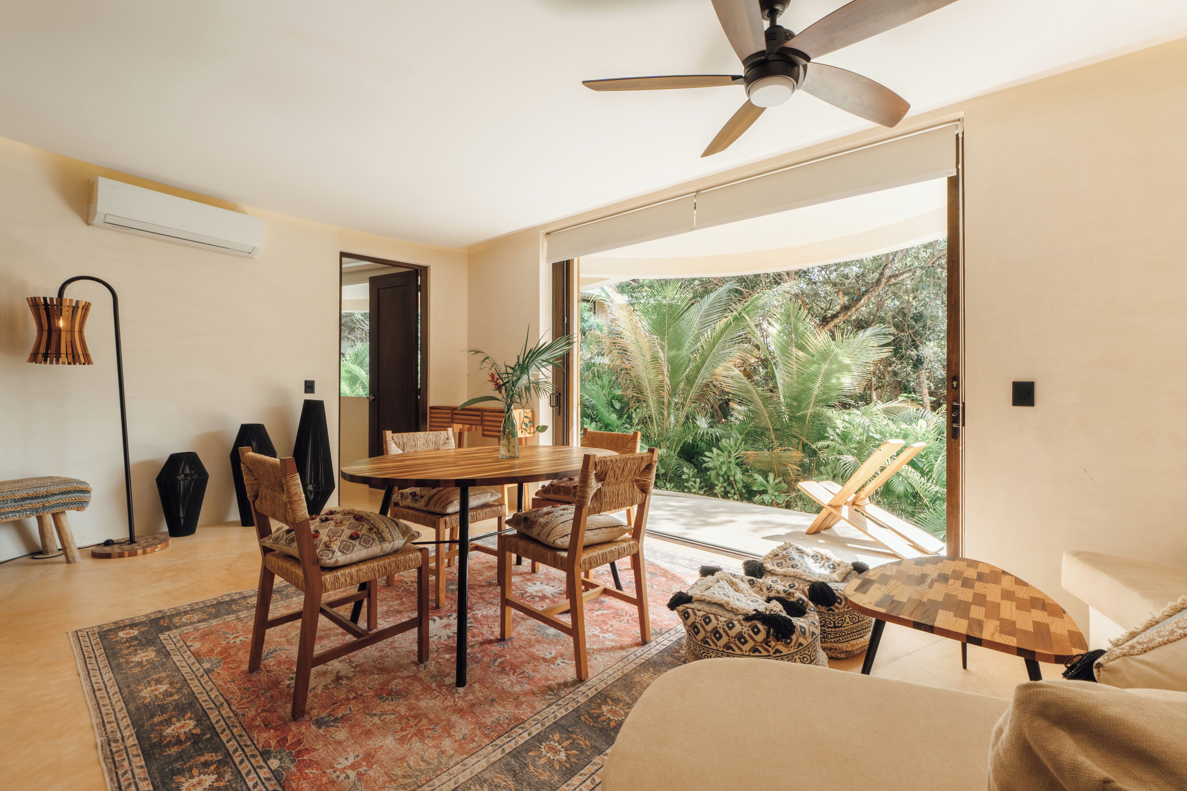 Une pièce spacieuse comprenant un coin repas, un ventilateur de plafond, une grande fenêtre donnant sur un espace verdoyant et une porte-fenêtre coulissante qui mène à l’extérieur.
