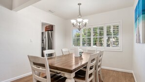 A dining area with a wooden table and chairs, a chandelier, a refrigerator, and a window with blinds. - Perfect Oasis w/ Pool, jacuzzi, close to airports, beach, entertainment & more! (Long Beach)