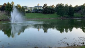 A tranquil pond with a fountain, surrounded by lush greenery and a building with a yellow facade in the background. - Bahama Bay Community, Pools, Basketball, Natural Trails and More! (Davenport)